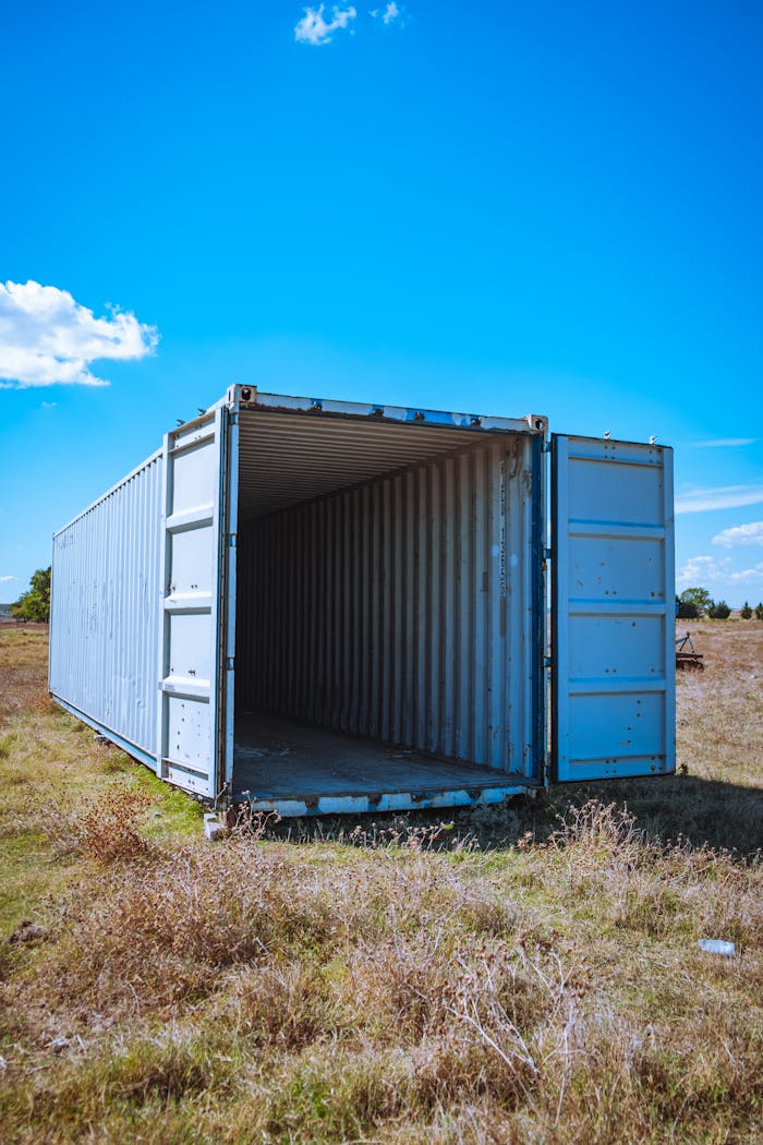 An open shipping container sits in an empty grassy field under a clear blue sky.